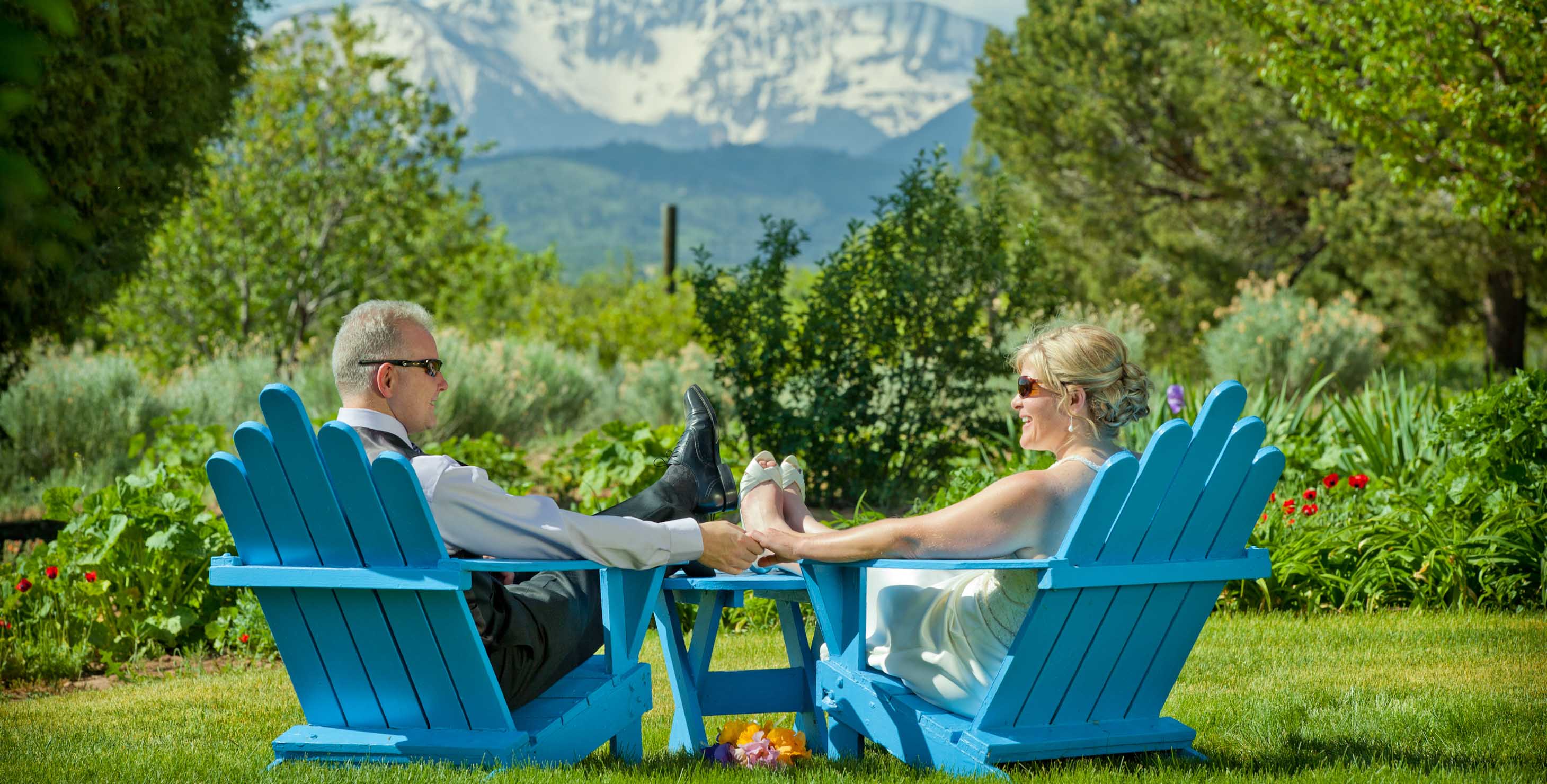 Elopement couple sitting on blue wooden chairs in a garden with mountains in the distance